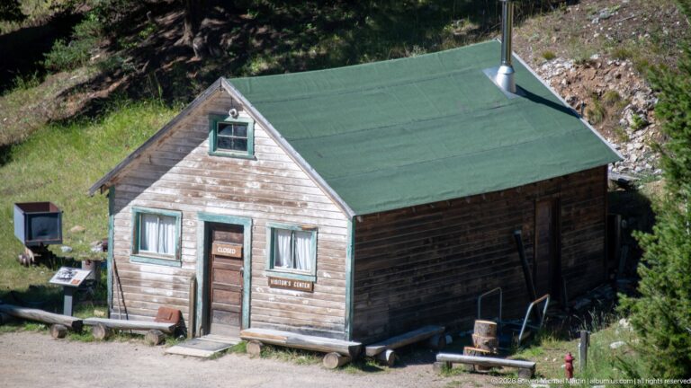 Weathered gable-front Garnet Visitor Center with green roof and trim, central door, two windows, and benches outside.