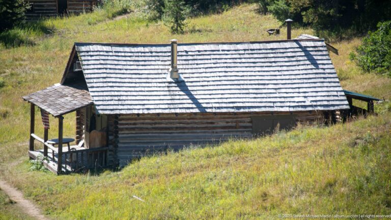Long log cabin with broad shingle roof, two stovepipes, small end porch, and side extension on a grassy hillside.