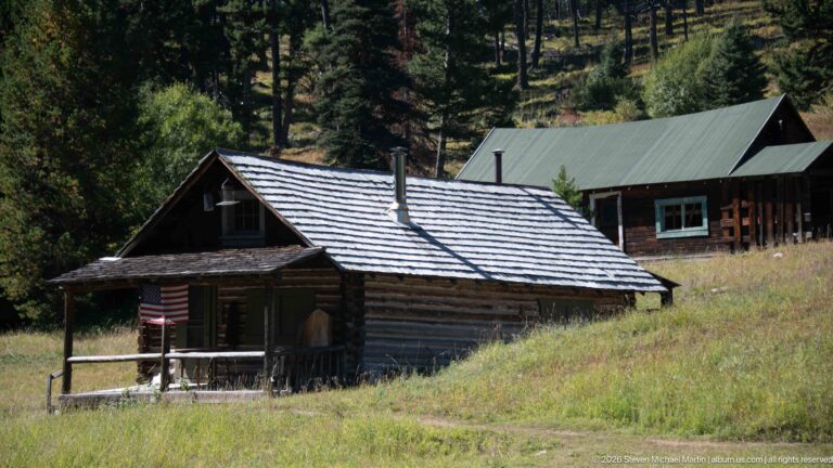 Small log cabin with porch and American flag on a grassy hillside, with a larger green-roofed log building behind.