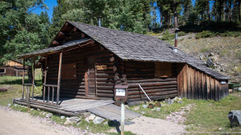 Small log cabin with shallow porch, wood-shingle roof, central door, side addition, and interpretive sign beside a gravel path.