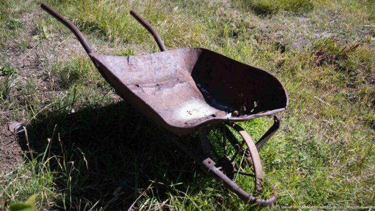 Rusting single-wheel metal wheelbarrow with long handles standing in sunlit grass at Garnet Ghost Town.