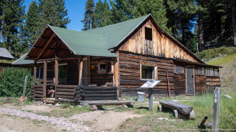 Porch-fronted log cabin with green gable roof, sawn-board gable, interpretive panel, and bench in a forest clearing.