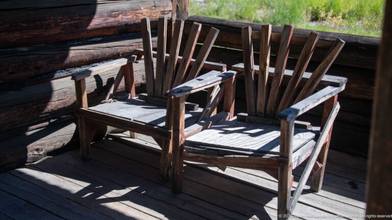 Two weathered wooden slatted armchairs side by side on a shaded porch with sunlight across their seats.