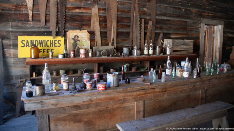 Long wooden bar lined with bottles, jars, tins, and cups beneath a yellow Sandwiches Coffee sign and large saw blades.