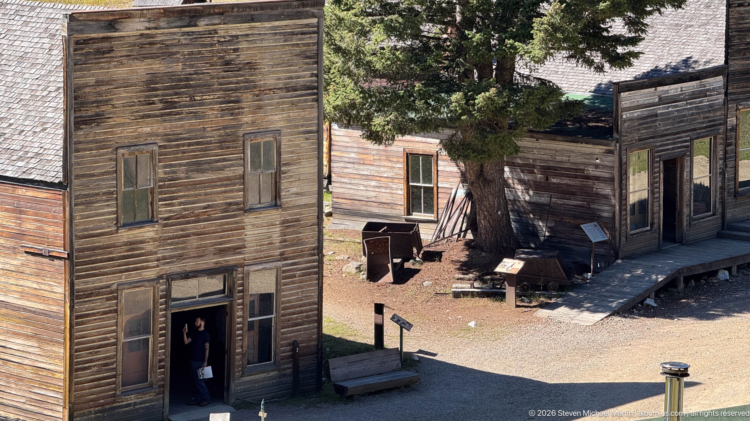 Tall weathered false-front building with a visitor in its doorway beside a lower storefront shaded by a large conifer.