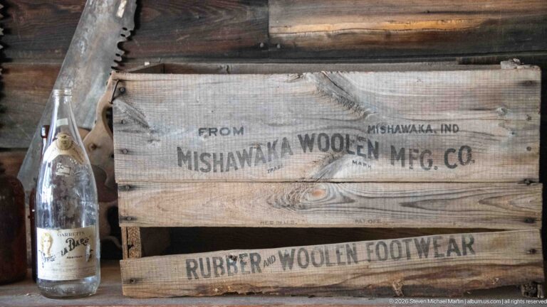 Weathered wooden crate stenciled Mishawaka Woolen Mfg. Co., Mishawaka, Indiana, and Rubber and Woolen Footwear beside a glass bottle.