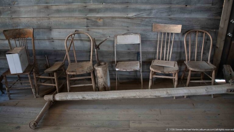 Row of mismatched wooden spindle-back and bentwood chairs with a metal folding chair against a plank wall.