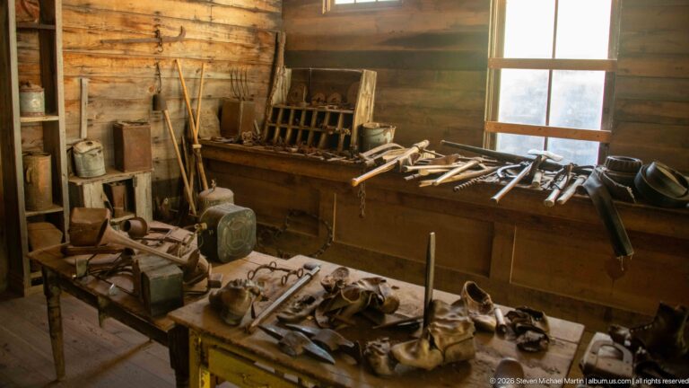 Workshop interior with worn shoes on a foreground table, tool-covered bench, shelves, chains, and long-handled implements.