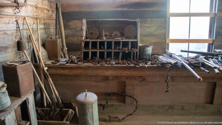 Workbench beneath a bright window covered with metal tools and hardware, with long-handled implements leaning against the log wall.