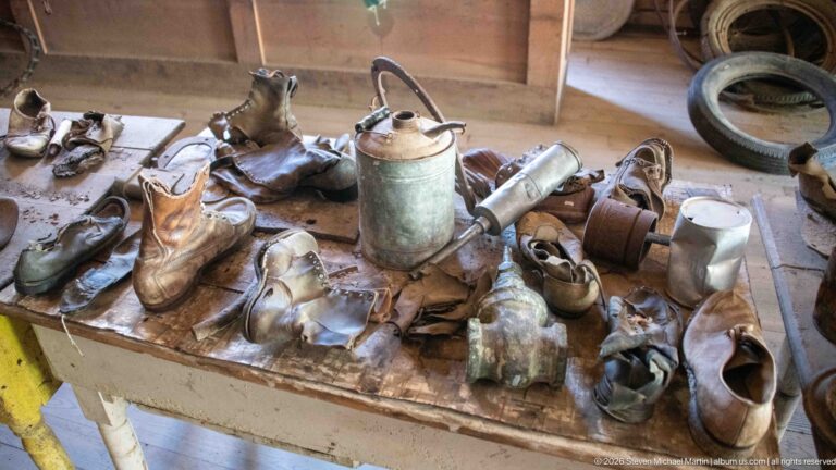 Table crowded with worn leather shoes and boots of different sizes beside old metal vessels and containers.