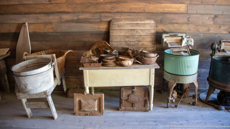 Galvanized wash tubs, pale work cabinet, cylindrical wringer washer, and old household appliances arranged against a log wall.