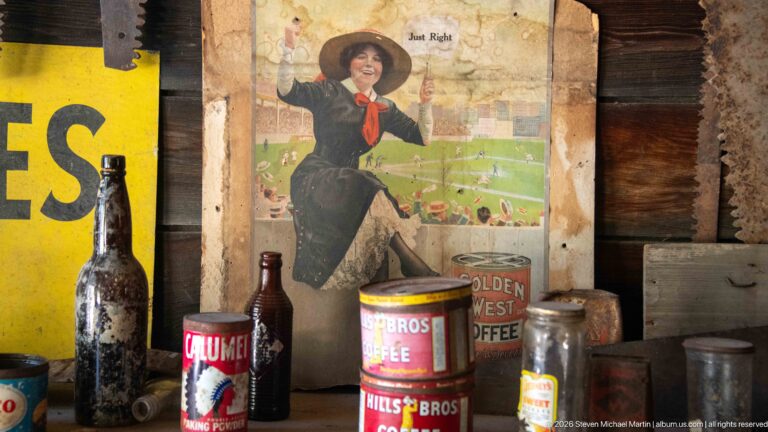 Historic coffee tins, bottles, and jars arranged before a colorful advertising print of a woman in a hat.
