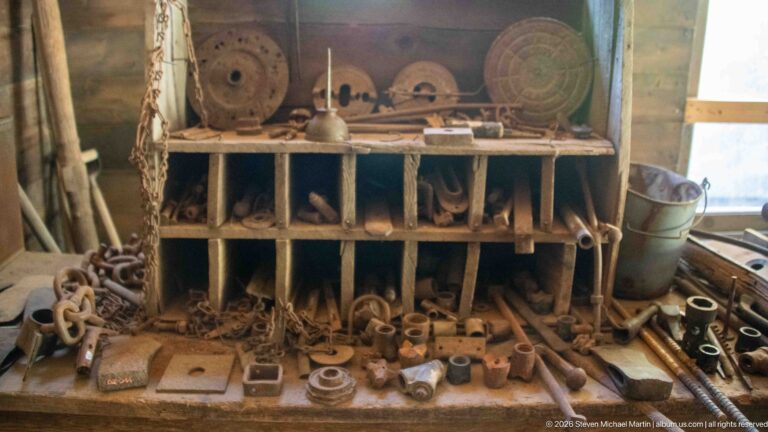 Wooden cubby organizer and workbench densely covered with chains, pulleys, bearings, fittings, and assorted metal hardware.