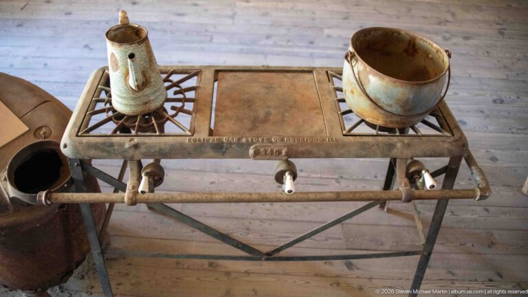 Three-burner metal cooking stove on slender legs with two old vessels resting on its grate and three front control valves.