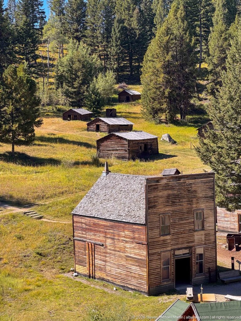 Elevated view of a two-story false-front building with smaller cabins scattered across a sunny grassy hillside among conifers.