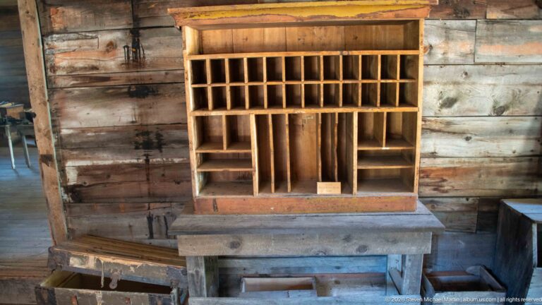Wooden pigeonhole organizer with many small compartments standing on a heavy workbench against weathered plank walls.