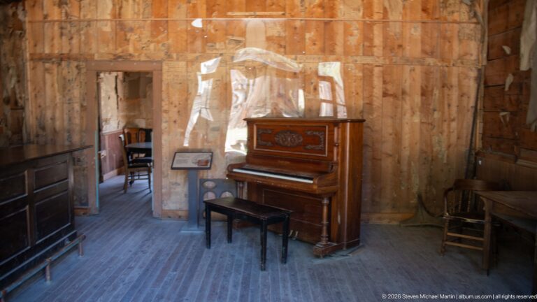 Decorated upright piano with bench against a newspaper-covered wooden wall, with chairs and an interpretive stand nearby.