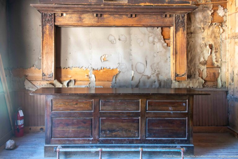 Long dark wooden bar with recessed panels standing against a heavily deteriorated wall in a plank-floored room.