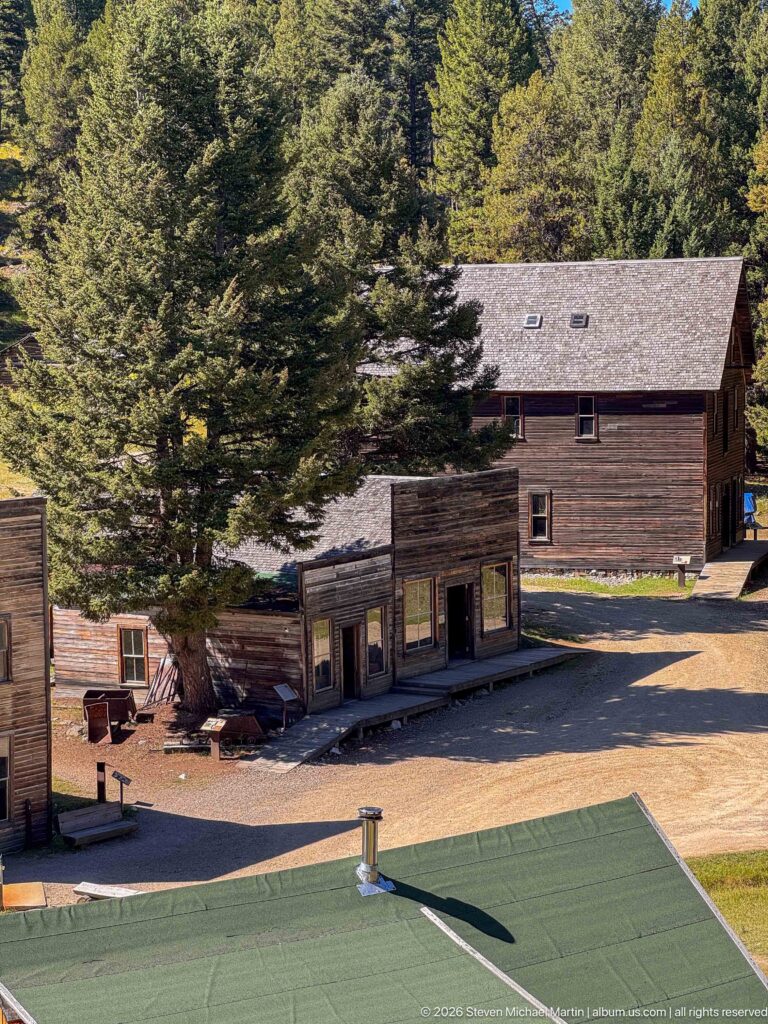 Elevated view over Garnet Ghost Town showing false-front buildings, a larger two-story structure, dirt street, green roof, and surrounding conifers.