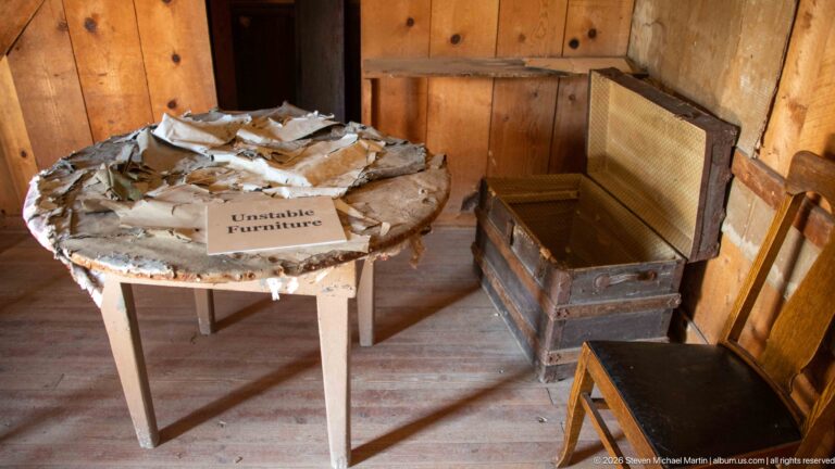 Damaged round table marked Unstable Furniture beside an open wooden chest and several chairs in a plank-walled room.