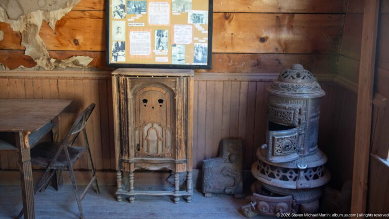 Ornate narrow wooden cabinet beside a decorative cast-iron stove, folding table, and chair against wood-paneled walls.