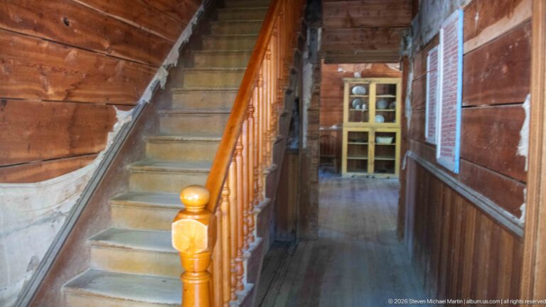 Wooden staircase with polished newel post and turned balusters beside a corridor leading toward a yellow cupboard.