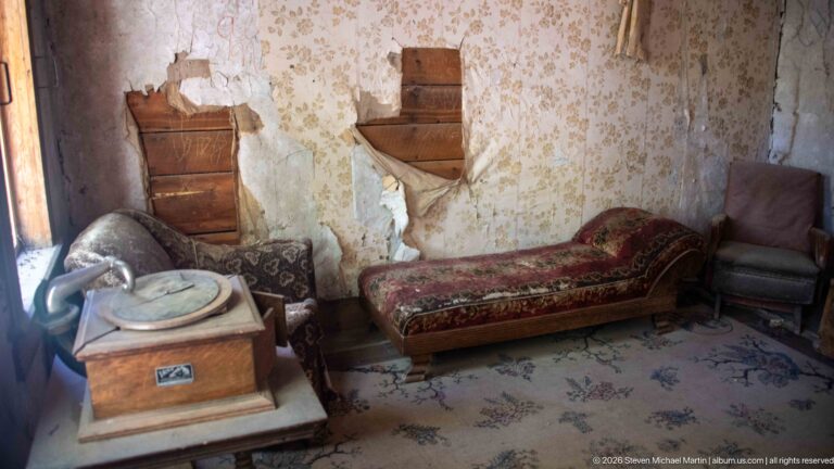 Floral chaise lounge beside an upholstered chair and cabinet phonograph in a room with torn patterned wallpaper and a floral floor covering.