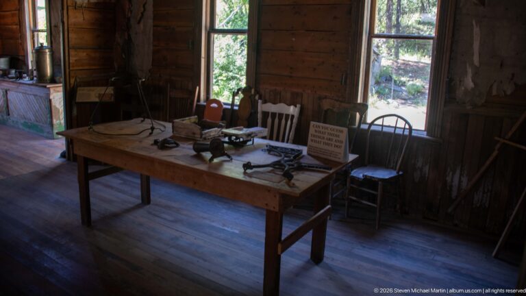 Wooden table displaying small historic tools and objects between two tall windows, with chairs and a do-not-touch sign.