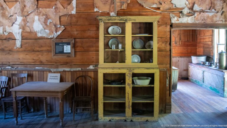 Yellow glass-front cupboard holding dishes beside a wooden table and chairs, with a green kitchen worktable visible through the doorway.