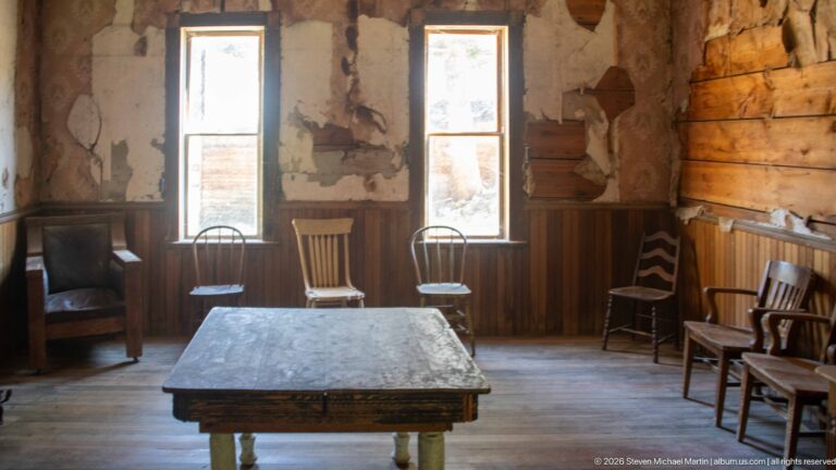 Square wooden table surrounded by mismatched chairs and rocking chairs between two tall windows in a deteriorated room.